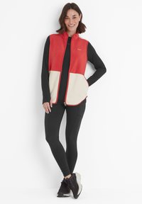 Red and cream fleece vest with a front zipper, two side pockets, and a high collar. Worn over a black long-sleeve top and leggings.