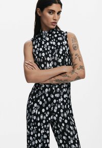 Svart ärmlös jumpsuit med stora silverfärgade prickar, med en hög krage och en slät, glänsande yta.