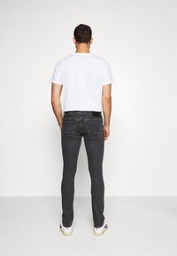 Man som bär en vit t-shirt, mörkgrå tajta jeans och vita sneakers, står vänd mot en enfärgad vit bakgrund.