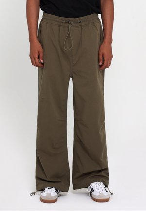 Persona che indossa pantaloni marroni larghi con vita con coulisse e sneakers bianche con strisce nere, in piedi contro uno sfondo semplice.