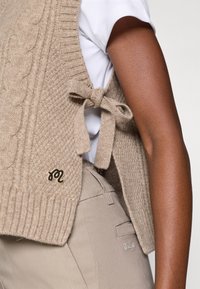 Vestă tricotată bej cu model cu torsade, legată lateral cu o fundă. Prezintă un mic logo negru. Purtați peste o cămașă albă.