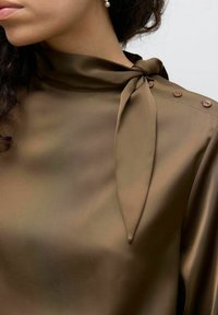 Haut en satin marron avec un col noué, présentant un léger éclat, une texture lisse et des boutons sur l'épaule.