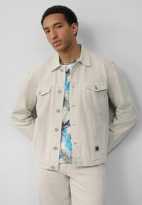 Beige denimjack met dubbele borstzakken en zilveren knopen; gedragen over een kleurrijk shirt met tropische print, gestyled met lichtgekleurde broek.