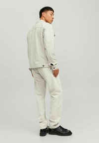 Giacca beige chiaro con una vestibilità leggermente ampia, bottoni neri e tasche frontali, abbinata a jeans bianco sporco e sneakers nere.