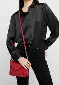 Donna che indossa una blusa di raso nera e pantaloni neri, mentre tiene una piccola borsa a tracolla rossa con catena dorata e il logo DKNY.