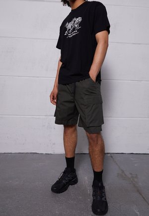 Mann trägt schwarzes T-Shirt mit Grafik, dunkelgrüne Cargo-Shorts, schwarze Socken und schwarze Sportschuhe, steht vor einer grauen Wand.