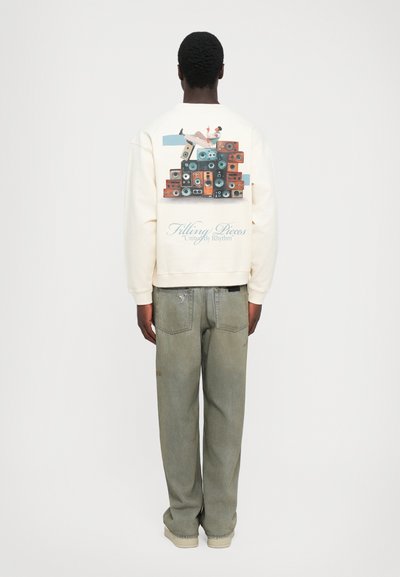 Homem com um sweatshirt creme com uma imagem de uma pessoa reclinada em colunas vintage empilhadas e o texto "Filling Pieces United By Rhythm" nas costas.