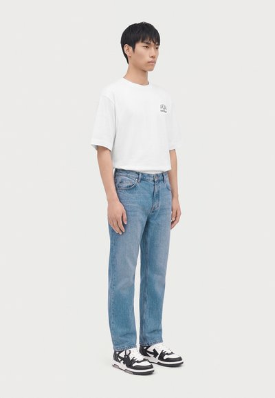 Jeune homme debout portant un t-shirt blanc, un jean bleu et des baskets noires et blanches sur un fond clair uni.