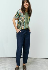 Chemise à manches courtes, colorée avec des motifs floraux verts, bleus et oranges, associée à un pantalon en denim bleu foncé à coupe décontractée et des chaussures marron à enfiler.