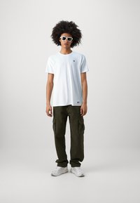 Maglietta bianca in cotone con un piccolo logo, pantaloni cargo verde oliva con tasche laterali e scarpe da ginnastica bianche, su uno sfondo neutro.