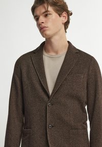 Braun gemusterte Blazer aus strukturiertem Stoff mit zwei Knöpfen und einer Brusttasche, darüber ein beiges T-Shirt.
