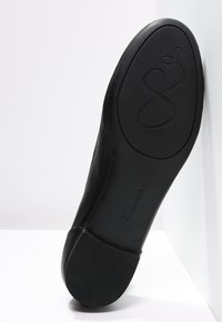 Semelle plate en cuir noir avec un talon et une zone de la plante texturés, présentant un motif décoratif en relief et le nom de la marque Clarks.