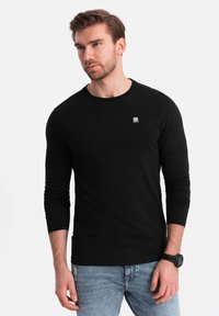 Schwarzes Langarmshirt mit Rundhalsausschnitt, aus glattem Baumwollstoff gefertigt. Verfügt über ein kleines Logo auf der Brust, kombiniert mit blauen Jeans.