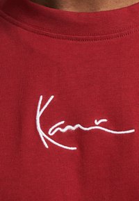 Rød bomulds-T-shirt med et hvidt broderet signaturdesign på forsiden, rund halsudskæring og blød tekstur.
