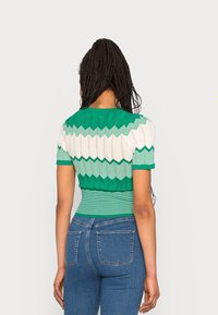 Grön och vit mönstrad kortärmad topp med ett zigzag-mönster och randig midja, kombinerad med blå denimjeans. Strukturera stickad kvalitet.