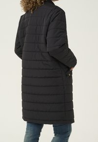 Abrigo tipo puffer negro con textura acolchada vertical, cuello acanalado y detalles de botones a presión. Diseño largo que llega a la mitad del muslo para mayor calidez.