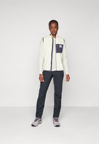 Veste en polaire pour femmes en blanc cassé avec un col montant, des accents navy et une poche poitrine zippée. Associée à un pantalon foncé et des chaussures de randonnée claires.