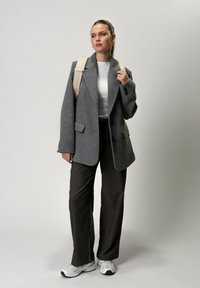 Blazer en mélange de laine gris, haut ajusté blanc, pantalon foncé et baskets blanches. Le modèle porte un sac à dos beige, avec une texture lisse et un design structuré.