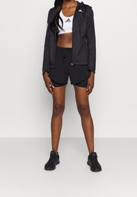 Nainen, joka on pukeutunut mustaan Adidas-takkiin, mustiin kerroksellisiin shortseihin, valkoiseen Adidas-urheiluliiviin ja mustiin lenkkareihin, seisoo valkoista taustaa vasten.