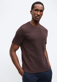 Άνδρας με κοντά σγουρά μαλλιά, φοράει υφασμάτινο σκούρου καφέ κοντομάνικο πουκάμισο και σκούρο μπλε παντελόνι, με τα χέρια στις τσέπες, ουδέτερο φόντο.