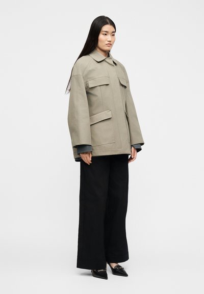 Γυναίκα που φοράει υπερμεγέθη μπεζ σακάκι με τσέπες με καπάκι, μαύρο παντελόνι με φαρδιά μπατζάκια και μαύρες γόβες με μυτερή μύτη, στέκεται μπροστά σε απλό φόντο.