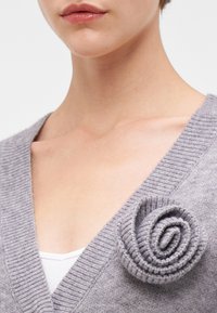 Harmaa neulotun neuletakin, jossa on V-pääntie, ribbikuviointi ja vasemmalla puolella ruusun muotoinen koristelu. Pehmeä ja kevyt materiaali.