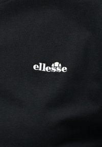 Μαύρο ύφασμα με μικρό λευκό κεντημένο λογότυπο της μάρκας "ellesse" με στυλιζαρισμένο σχέδιο ημικυκλίου πάνω από τα γράμματα.
