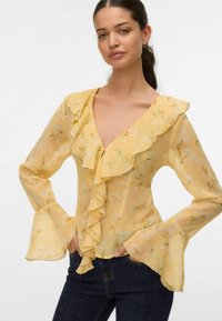 Blouse florale jaune avec un col froncé et des manches évasées. Tissu transparent avec un léger motif floral, présentant un décolleté en V profond et une texture douce.