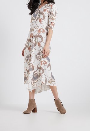 Femme portant une robe midi blanche à imprimé cachemire et des bottines beiges en daim, debout devant un fond uni.