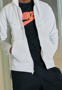 Biała bluza z zamkiem błyskawicznym z białymi kieszeniami bocznymi i lekkim połyskiem, noszona na czarnej koszulce z wyraźnym pomarańczowym logo Nike, do tego ciemne spodnie.