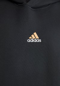 Črna kapuca iz mehkega blaga slog, ki vsebuje Adidasov logotip v zlati in beli barvi, s klasičnim dizajnom ter sprednjim kenguru žepom.