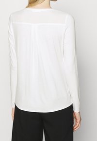 Biała bluzka z długim rękawem i okrągłym dekoltem, z pionowym szwem biegnącym wzdłuż pleców. Gładka tekstura, luźny krój.