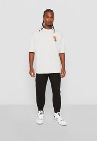 Licht beige oversized t-shirt met kleurrijke vlamgrafiek, gecombineerd met zwarte cargo broeken en witte high-top sneakers.