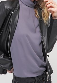 Giacca di pelle nera sopra un top a collo alto viola. L'outfit presenta una texture liscia, un design aderente e una chiusura con zip.