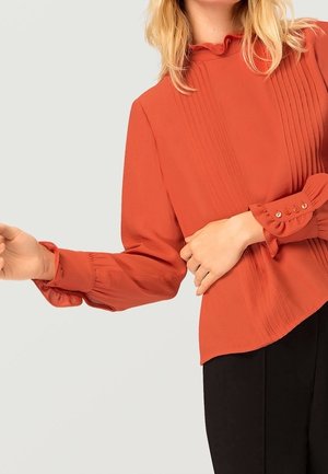 Vrouw draagt een blouse met lange mouwen in verbrande oranje kleur met plooien en knoopmanchetten, gecombineerd met een zwarte broek tegen een effen achtergrond.