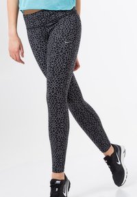 Leggings neri con un motivo geometrico bianco, caratterizzati da un alto fascione in vita e un tessuto elasticizzato. Indossati con scarpe da ginnastica nere.
