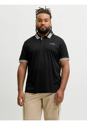 Čovjek s dredovima u crnoj polo majici s bijelim detaljima i bež cargo kratkim hlačama, stoji na jednobojnoj svijetloj pozadini.