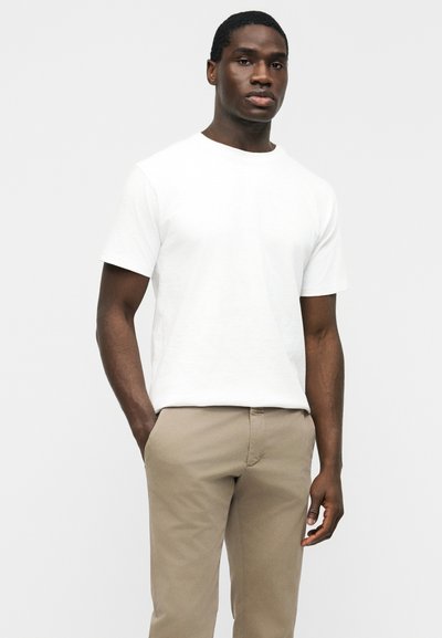 Homme debout, les mains dans les poches, portant un t-shirt blanc uni à manches courtes et un pantalon beige, sur un fond neutre.