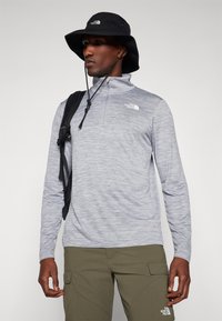 Lys grå, langermet zip-up skjorte med en jevn tekstur, sammen med olivenfargede bukser og en svart bøttehatt. Svart ryggsekk inkludert.