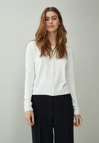Giovane donna con capelli lunghi e mossi che indossa un cardigan bianco con bottoni e pantaloni neri, in piedi davanti a uno sfondo grigio chiaro uniforme.