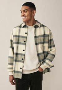 Plaid flanellsärk kreemja ja rohelise tooni ning mustade nööpidega, millel on rinnatasku ja üles keeratud varrukad, kantud heleda dressipluusi üle.