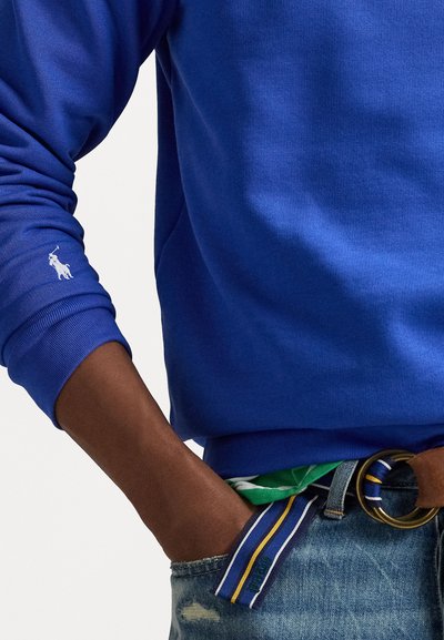 Karaliski zila sporta krekls ar ribotām piedurknēm, uz kuras ir balta polo spēlētāja logo uz piedurknes. Savienots ar bālām džinsām un daudzkrāsainu jostu.