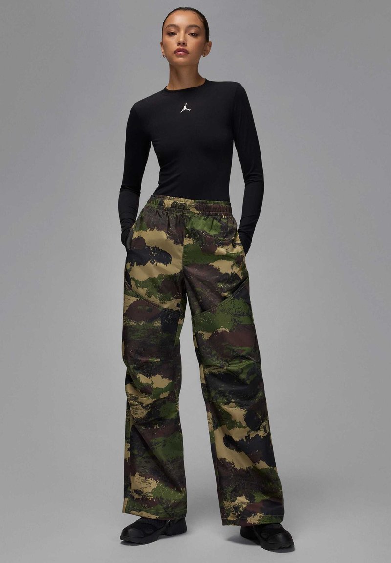 Haut noir à manches longues avec logo blanc, associé à un pantalon large en camouflage vert et marron avec des motifs texturés, et des chaussures noires.