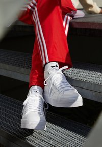 Person wearing white Adidas high-top sneakers and red track pants with white stripes sitting on metal stairs.