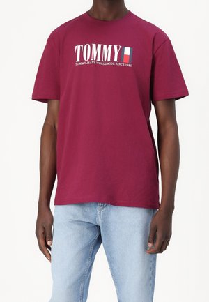 Muž v bordó tričku Tommy Jeans s logem a světle modrých džínách stojí před jednobarevným bílým pozadím.