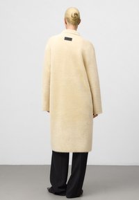 Blonde vrouw met haar in een knot, draagt een lange crèmekleurige fuzzy jas en zwarte wijde broek, staat met de rug naar ons toe op een grijze vloer.