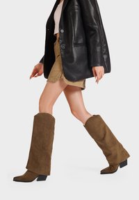 Stivali marroni in suede al ginocchio con punta quadrata e tacco impilato, abbinati a un blazer in pelle nera e pantaloni corti color khaki.