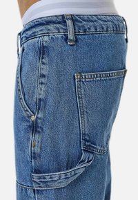 Siniset farkut varustettuina teksturoidulla pinnalla, sivutaskulla ja näkyvillä sauman yksityiskohdilla. Vaikutelma kestävästä ja rennosta mallista.