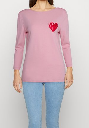 Femme portant une chemise à manches longues rose clair avec un motif de cœur rouge sur le côté gauche de la poitrine, associée à un jean bleu clair.