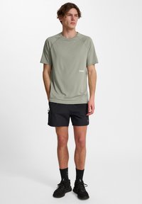 Maglietta atletica a maniche corte di un leggero verde oliva, con maniche raglan, caratterizzata da un piccolo logo bianco sul lato sinistro. Indossata con pantaloni corti neri.